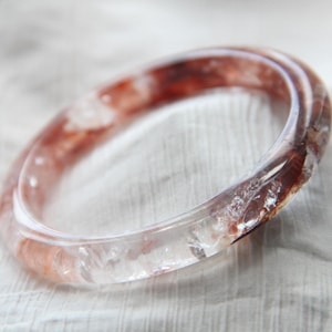 May include: A clear quartz bracelet with reddish-brown inclusions. The transparent bangle is circular and rests on a white, textured surface. The gemstone bracelet is a piece of jewellery.