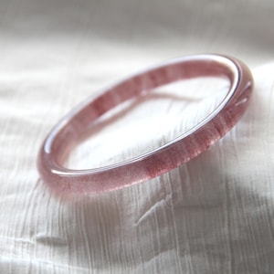 May include: A polished, circular bracelet made from a translucent, pinkish-red gemstone. The bangle's surface is smooth, revealing a natural pattern within the stone. The bracelet is displayed against a soft, white backdrop.