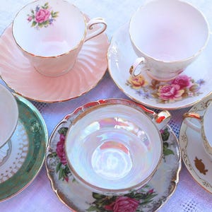May include: A collection of vintage teacups and saucers. The cups and saucers are in various colors, including pink, white, and green, and feature floral designs and gold trim. The teacups are arranged on a white surface.