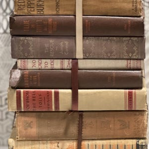 May include: A stack of vintage books bound together with a cream-colored ribbon and a dark brown leather strap. The books have various shades of brown covers, with titles like "Red Pepper Burns" and "For Whom the Bell Tolls" visible.