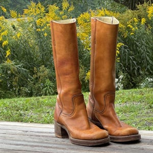 May include: A pair of knee-high tan leather boots featuring a structured shaft, a rounded toe, and a stacked block heel. The boots have visible tonal stitching along the sides and a smooth finish, showcasing a classic pull-on design with a sturdy sole.