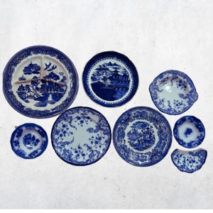 May include: A collection of blue and white ceramic plates with various patterns. The plates include a divided plate, a small bowl, and several round plates with floral and landscape designs. The collection is arranged on a white surface.