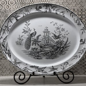 May include: Oval serving platter with a black and white peacock and pagoda design. The rim features a floral and bird pattern. The platter is displayed on a black metal stand. A decorative piece for serving food or as a display item.