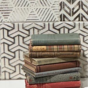 May include: A stack of antique books with aged covers in various colors, including green, red, and brown. The books are stacked on a white surface, with a patterned wall in the background.
