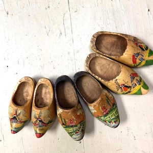 May include: Five wooden clogs arranged in a group, each featuring hand-painted scenes of windmills, houses, and landscapes in yellow, red, green, and blue tones. The shoes have a natural wood finish and include the word Holland painted on them.
