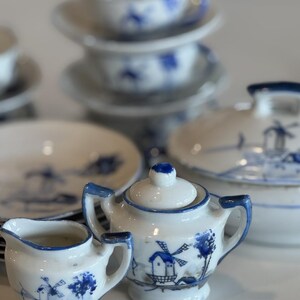 May include: A collection of white porcelain tea set pieces with blue windmill and tree designs. The set includes a creamer, sugar bowl, cups, saucers, and a serving dish. The rims and handles are accented with blue paint.