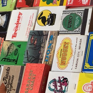 May include: A collection of vintage matchbooks with various designs and text. The matchbooks feature logos and names of hotels, casinos, and restaurants, including "Treasury," "Carson Nugget," and "Marrakech." The colors are diverse, with red, green, yellow, and blue.