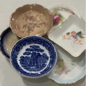 May include: A collection of small, decorative vintage dishes. The dishes feature floral and landscape designs in various colors, including blue, gold, and pink. The shapes vary from round to square, with scalloped and gilded edges.