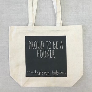 Puede incluir: Una bolsa de lona blanca con asas negras. La bolsa tiene un rectángulo negro con texto blanco que dice "PROUD TO BE A HOOKER" y una dirección de sitio web debajo.