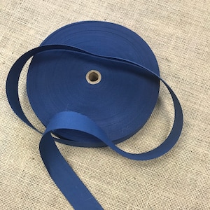 Puede incluir: Un rollo de cinta de sarga de algodón azul oscuro. La cinta está enrollada y un trozo de cinta cuelga.