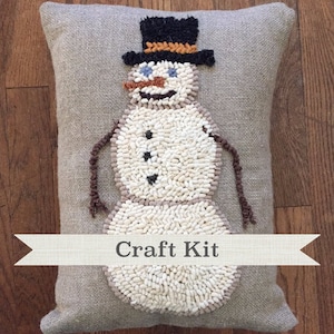 Peut inclure: Un coussin rectangulaire en toile de jute avec un motif de bonhomme de neige. Le bonhomme de neige est fait de tissu texturé blanc et crème, avec un chapeau haut de forme noir, des yeux bleus et un nez en carotte marron. Le texte "Craft Kit" est affiché sur une bannière.