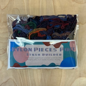 May include: A clear plastic bag filled with a variety of hand-dyed nylon yarn scraps in shades of blue, green, red, and brown. The bag has a label that reads "Nylon Pieces Pack Stash Builder 2oz mixed hand dyed colored nylon bundle. Weights vary from bulky to extra chunky."