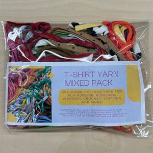 May include: A clear plastic bag filled with colorful t-shirt yarn. The yarn is in various colors and patterns, including red, blue, green, yellow, and white. The bag also includes a label that reads "T-Shirt Yarn Mixed Pack 402 Mixed Pattern Yarn for Rug Hooking, Punching, Weaving, Crochet, Knitting and More! Ecotextiles Recycled Yarn Ranging in Size from 1-3 inches. Curled to make Super Bulky Yarn Comparable to #5- Rug Hooking Wools. 85% Cotton Includes a mix of pieces that are 3-5 feet long."
