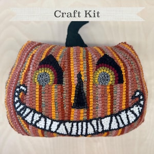 May include: Craft kit to make a grinning pumpkin pillow. The pillow is made of orange, red, and yellow striped fabric with black eyes and a wide, toothy grin.