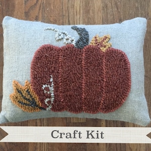 May include: Craft kit featuring a gray pillow with a brown, orange, and yellow felt pumpkin design. The pillow has the text "Craft Kit" printed on a white ribbon.