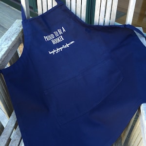 Puede incluir: Un delantal azul oscuro con un bolsillo blanco y el texto "Proud to be a Hooker" y "loopyloudesigns.com" bordado en la parte delantera.