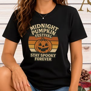 以下が含まれることがあります： ミッドナイトパンプキンフェスティバルのレトロなデザインが施された黒いTシャツ。デザインには、笑顔のカボチャと「Stay Spooky Forever」のテキストがヴィンテージスタイルで描かれています。Tシャツはクルーネックです。