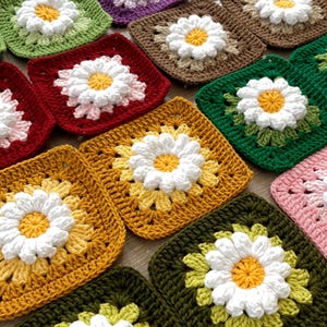 Op de afbeelding: Een verzameling kleurrijke, gehaakte madeliefjesvierkanten. Elk vierkant heeft een witte madelief met een gele kern, omringd door bloemblaadjes in verschillende kleuren zoals groen, geel, roze en bordeauxrood. De vierkanten zijn dicht bij elkaar geplaatst en tonen het handgemaakte ambacht.