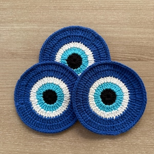 Puede incluir: Tres posavasos azules de ganchillo con un diseño de ojo turco. Cada posavasos presenta un círculo negro central, rodeado de anillos blancos, celeste y azul oscuro. Los posavasos están dispuestos sobre una superficie de madera marrón claro.