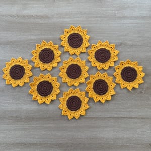 May include: A collection of ten handmade crochet sunflower coasters. Each coaster features a brown center and yellow petals, arranged on a gray wooden surface. The coasters are designed to resemble sunflowers.