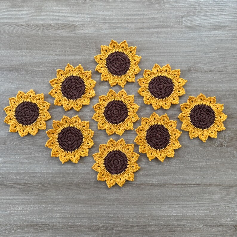 May include: A collection of ten handmade crochet sunflower coasters. Each coaster features a brown center and yellow petals, arranged on a gray wooden surface. The coasters are designed to resemble sunflowers.