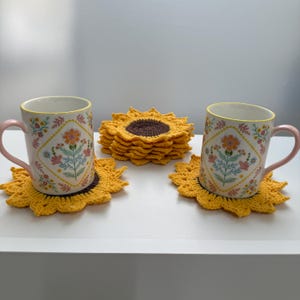 May include: Two ceramic mugs with floral patterns and pink handles sit on yellow crochet sunflower coasters. A stack of sunflower coasters is in the background. The mugs have a yellow rim.