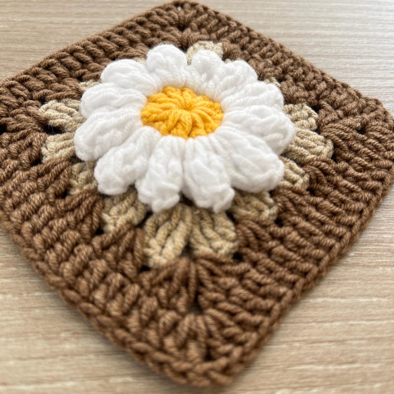 May include: A square, crocheted coaster with a daisy motif. The daisy has white petals and a yellow centre, set against a brown and beige background. The coaster is handmade and has a textured appearance.