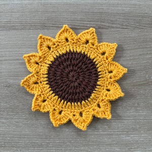 May include: A handmade crochet sunflower coaster. The coaster features a dark brown center and yellow petals. The petals have a scalloped edge. The coaster is round and sits on a wooden surface.