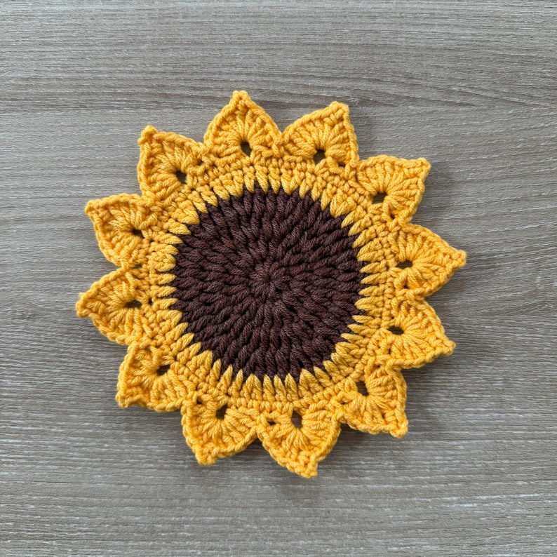 May include: A handmade crochet sunflower coaster. The coaster features a dark brown center and yellow petals. The petals have a scalloped edge. The coaster is round and sits on a wooden surface.