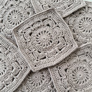 May include: Close-up of several crocheted squares in a neutral taupe color. Each square features a central floral design surrounded by intricate patterns. The squares are arranged in a slightly overlapping manner, showcasing the texture and detail of the handmade craft.