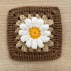 May include: A square, handmade crochet coaster with a daisy design. The daisy has white petals and a yellow centre, set against a beige and brown background. The coaster is made of yarn and has a textured appearance.