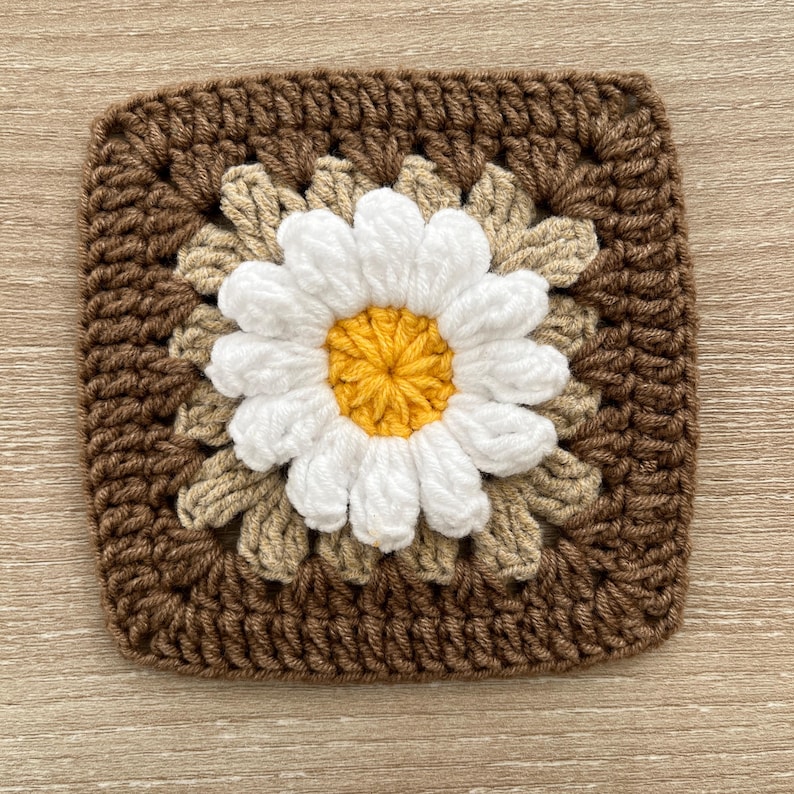 May include: A square, handmade crochet coaster with a daisy design. The daisy has white petals and a yellow centre, set against a beige and brown background. The coaster is made of yarn and has a textured appearance.