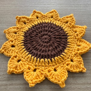 May include: A handmade crochet sunflower coaster. The coaster features a brown center and yellow petals. The petals are detailed with small holes and a scalloped edge. The coaster is designed to resemble a sunflower.