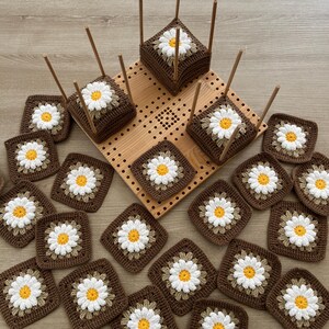 May include: Handmade crochet squares with daisy designs in brown, white, and yellow. Some squares are arranged on a wooden pegboard and wooden stands, showcasing a craft project. The squares are square-shaped.