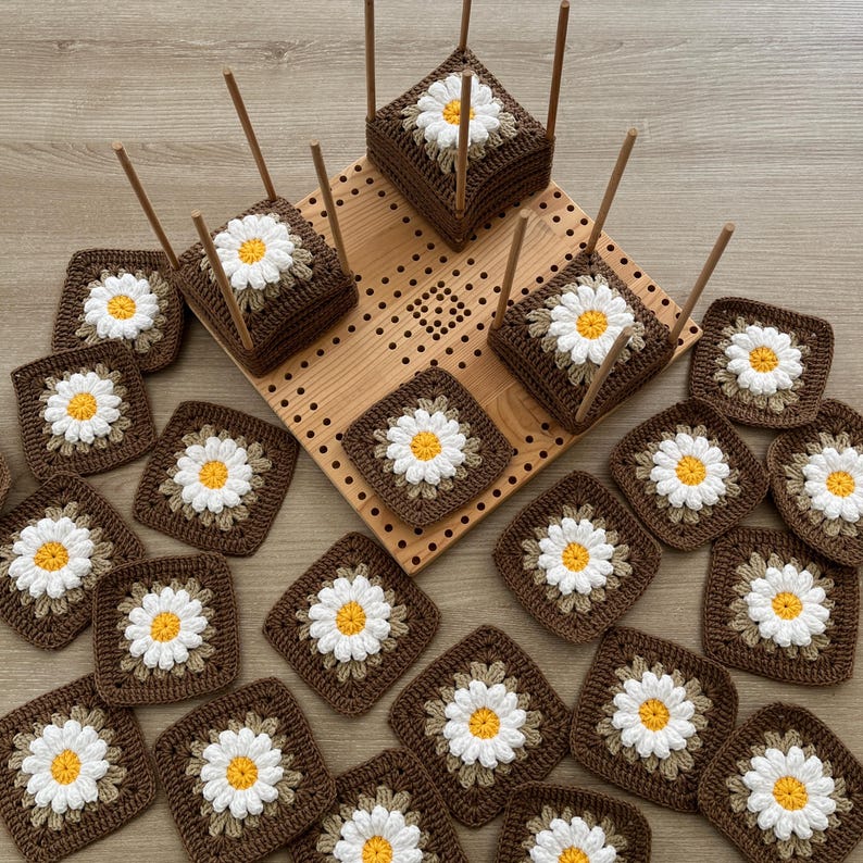 May include: Handmade crochet squares with daisy designs in brown, white, and yellow. Some squares are arranged on a wooden pegboard and wooden stands, showcasing a craft project. The squares are square-shaped.