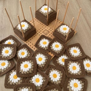 May include: A collection of handmade crochet daisy coasters in brown, white, and yellow. The square coasters, featuring a daisy design, are arranged on a wooden surface. Some are displayed on wooden stands with dowels, creating a unique presentation.