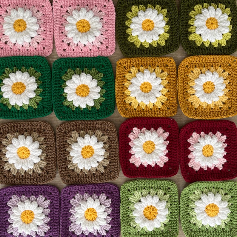 May include: A selection of crocheted squares in various colours, each with a daisy motif. The squares are in shades of pink, green, yellow, brown, red, purple, and green. The daisies have white petals and yellow centres.