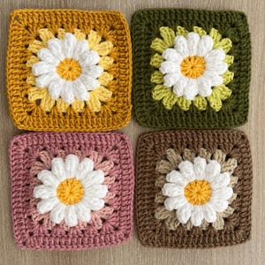 May include: Four crocheted daisy squares in shades of gold, green, pink, and brown. Each square features a white daisy with a yellow centre. The squares are handmade and have a textured appearance, ideal for craft projects.
