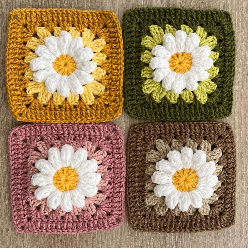 May include: Four crocheted daisy squares in shades of gold, green, pink, and brown. Each square features a white daisy with a yellow centre. The squares are handmade and have a textured appearance, ideal for craft projects.