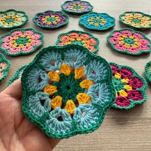May include: A selection of handmade crochet coasters in a floral design. The coasters are in various colours, including green, blue, yellow, pink, and purple. They are displayed on a light-coloured wooden surface.