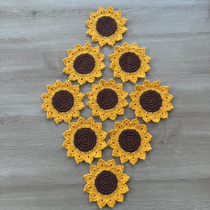 May include: A collection of ten handmade crochet sunflower coasters. Each coaster features a dark brown center and yellow petals, arranged in a diamond pattern on a wooden surface. The coasters are designed to protect surfaces from heat and moisture.