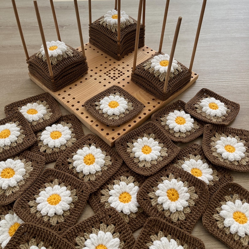 May include: Handmade crochet daisy squares in brown, white, and yellow. Some squares are stacked on a wooden pegboard with wooden dowels. The daisy squares are arranged on a light wood surface.