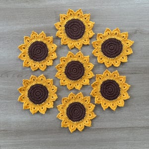 May include: Seven handmade crochet sunflower coasters. Each coaster features a brown center and yellow petals, resembling a sunflower. The coasters are arranged on a wooden surface, showcasing the intricate details of the crochet work.