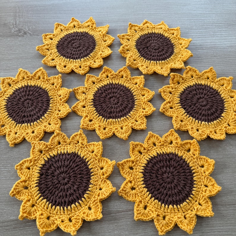May include: A collection of seven handmade crochet coasters. Each coaster is designed to resemble a sunflower, featuring a brown center and yellow petals. The coasters are arranged on a light-colored surface.
