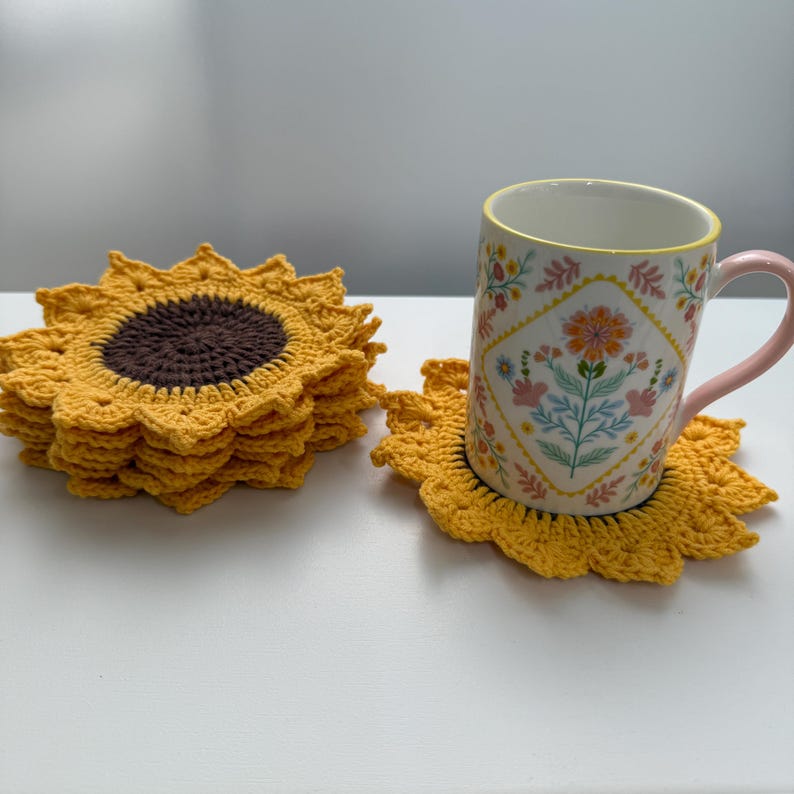 May include: A stack of yellow crochet sunflower coasters with a brown center. A floral patterned mug with a pink handle sits on one of the coasters. The coasters are handmade and add a touch of nature to any home decor.