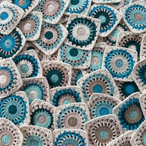 Könnte beinhalten: Eine Ansammlung handgefertigter Häkelquadrate in verschiedenen Blau-, Türkis- und Beigetönen. Jedes Quadrat weist ein kreisförmiges Design innerhalb eines quadratischen Rahmens auf, wodurch ein strukturierter, geschichteter Effekt entsteht. Die Quadrate sind eng angeordnet und zeigen die komplizierten Details der Garnarbeit.