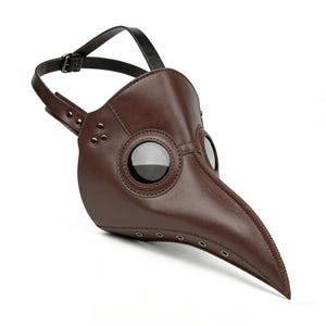 Op de afbeelding: Een bruin leren pestdoktermasker met een lange, snavelachtige neus en grote, ronde ooglenzen. Het masker heeft een zwarte riem om het op het hoofd te bevestigen en is gedetailleerd met klinknagels.