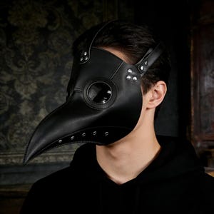 Op de afbeelding: Zwart leren pestdoktermasker met een lange snavel, ronde oogopeningen en geklonken details. Het masker heeft riemen om rond het hoofd te bevestigen. Een gotisch accessoire voor cosplay- of kostuumevenementen.