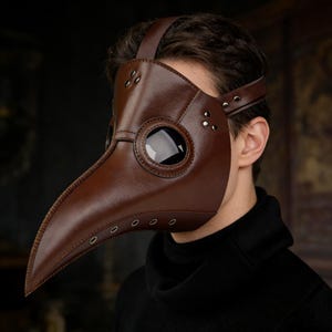 Op de afbeelding: Een bruin leren pestdoktermasker met een lange snavel en grote, ronde oogopeningen. Het masker heeft een band die over het hoofd gaat en is voorzien van klinknagels voor detail. Een historisch kostuumaccessoire.