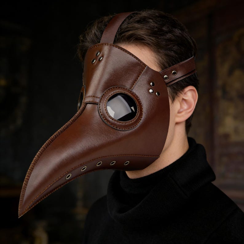Op de afbeelding: Een bruin leren pestdoktermasker met een lange snavel en grote, ronde oogopeningen. Het masker heeft een band die over het hoofd gaat en is voorzien van klinknagels voor detail. Een historisch kostuumaccessoire.